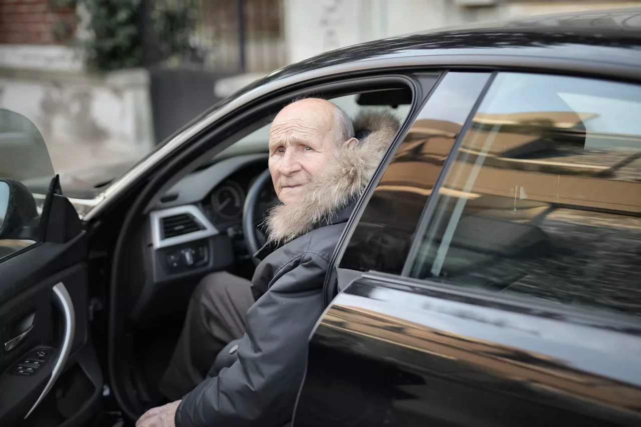 Ein älterer Mann in Jacke sitzt bei offener Tür im Auto und schaut heraus