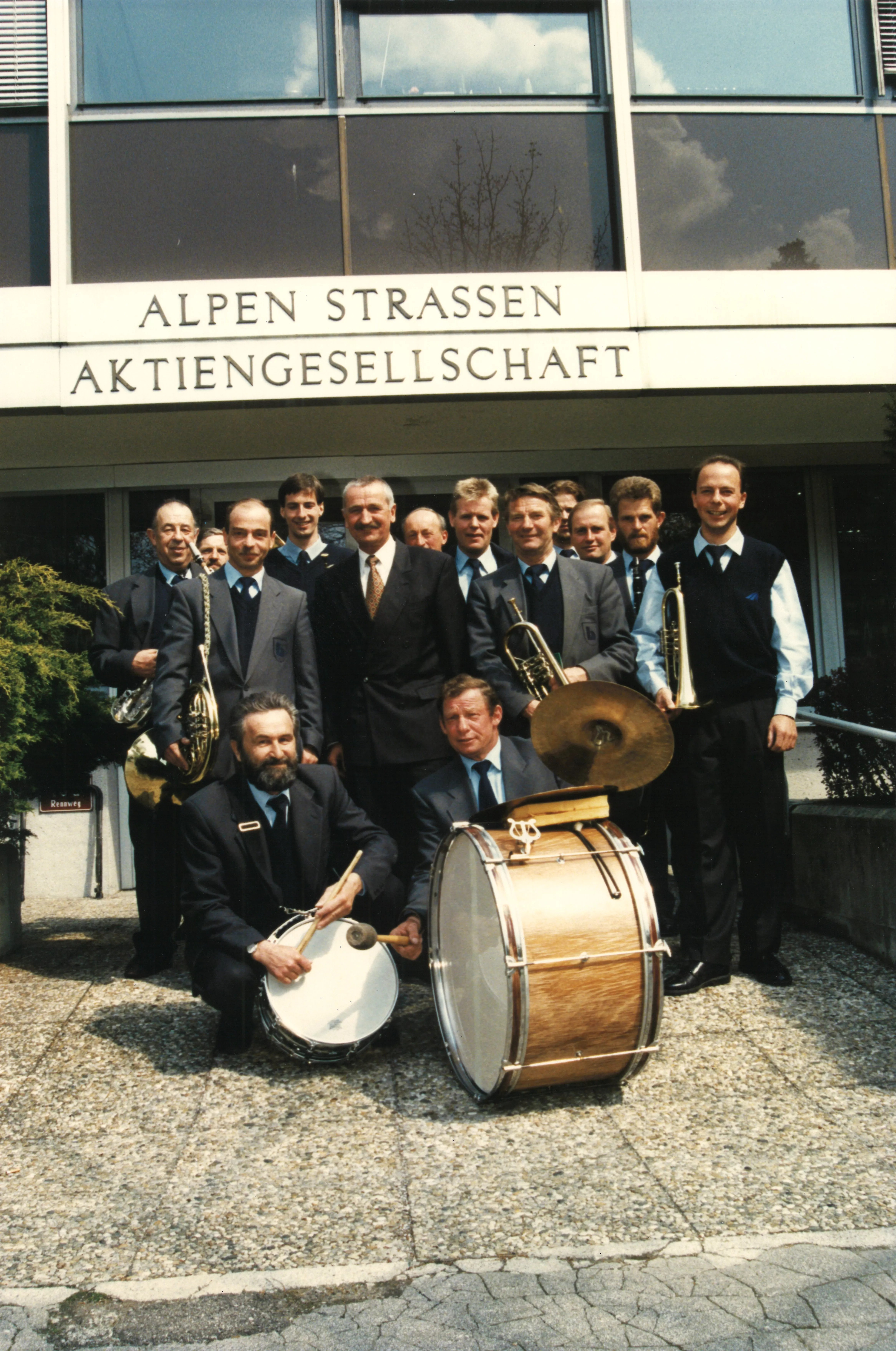 Eine Gruppe Männer in Anzügen steht mit Musikinstrumenten vor einem Gebäude