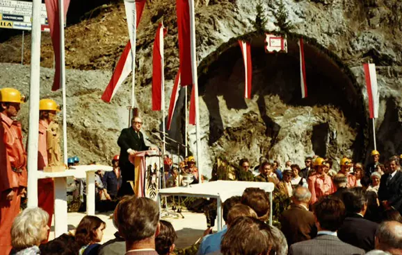 Eine Menschenmenge steht vor einem Podium, auf dem ein Mann vor einem Pult steht. Im Hintergrund ist das noch undurchbrochene Tunnelportal