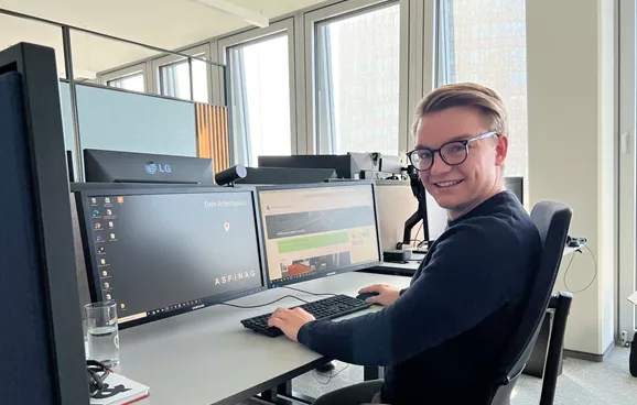 Ein junger Mann sitzt an einem Schreibtisch vor einem Computer und lächelt in die Kamera